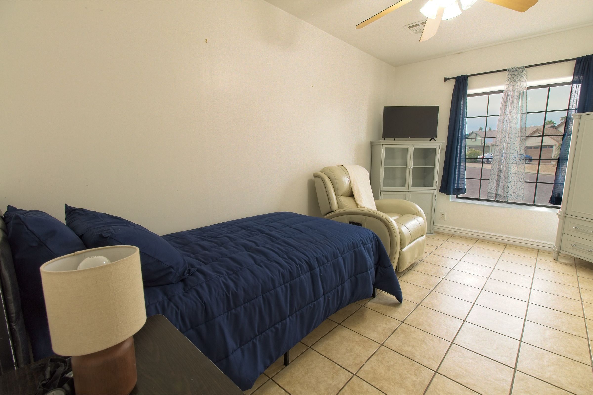 A bedroom with a navy bedspread, a cream recliner, a television and a window
