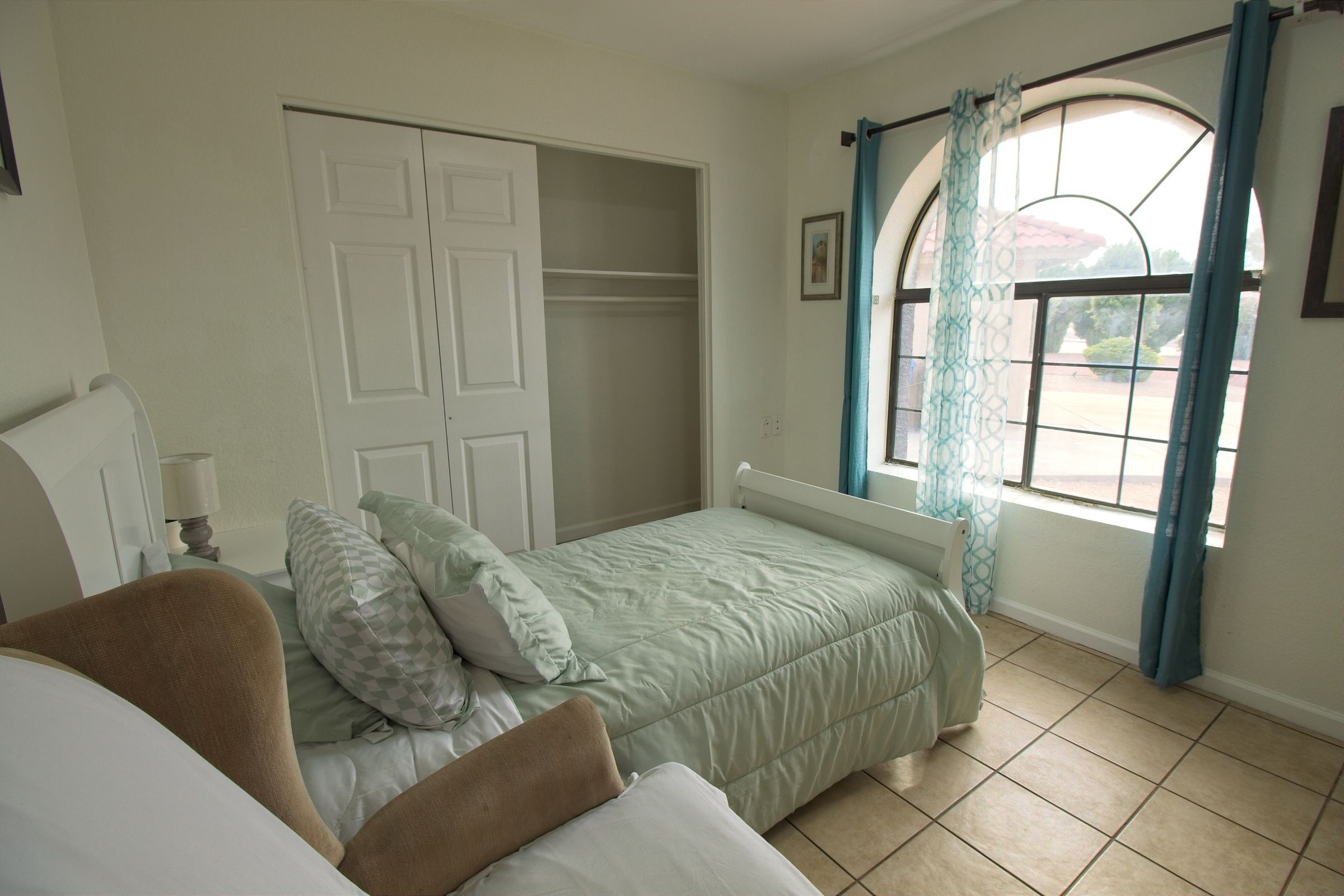 A private bedroom with sage bedding, a closet and an arched window with teal curtains